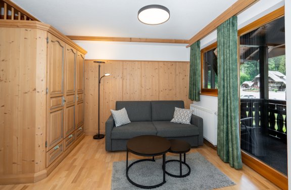 Wohnzimmer mit grauem Sofa, Holzwand, großem Schrank links und Balkon rechts mit grünen Vorhängen