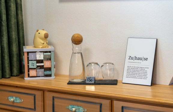 Holzkommode mit Spielzeug, Karaffe mit Holzdeckel, zwei Gläsern und Schild mit Text zu Zuhause